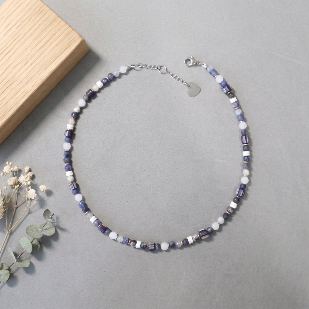 A beaded necklace with alternating lavender-blue and white round beads, accented by small silver-toned spacer beads and a silver heart charm, displayed on a gray surface next to a wooden block and dried flowers.