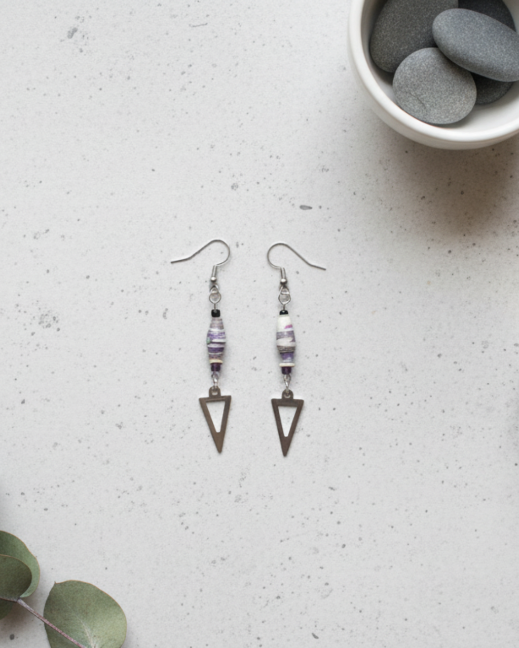 Pair of triangle-shaped earrings on a textured surface with a bowl of stones in the background.