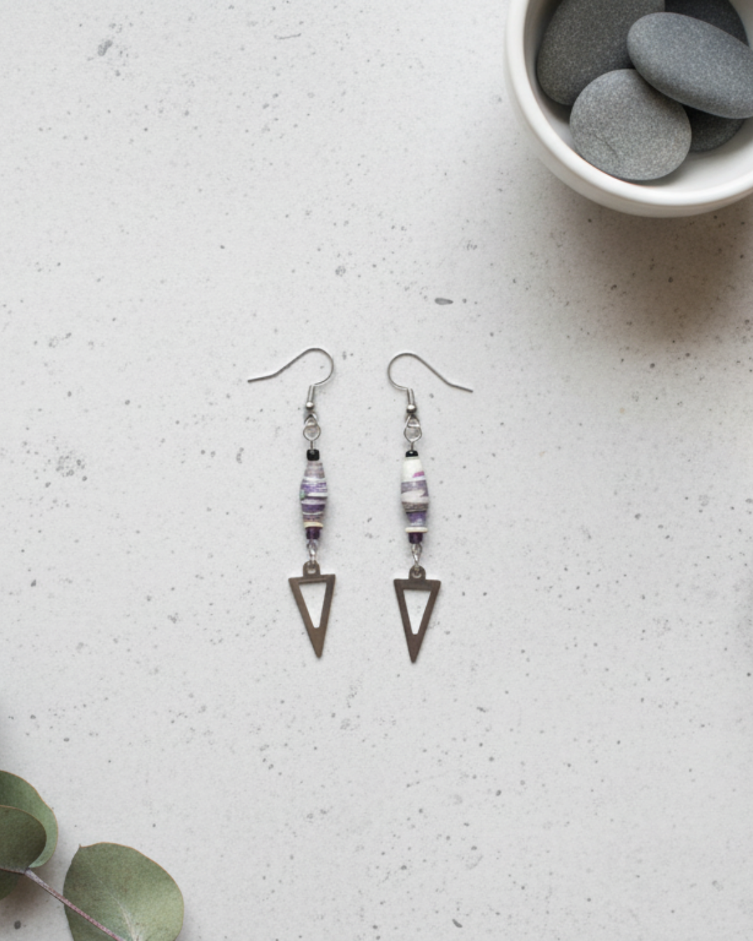 Pair of triangle-shaped earrings on a textured surface with a bowl of stones in the background.