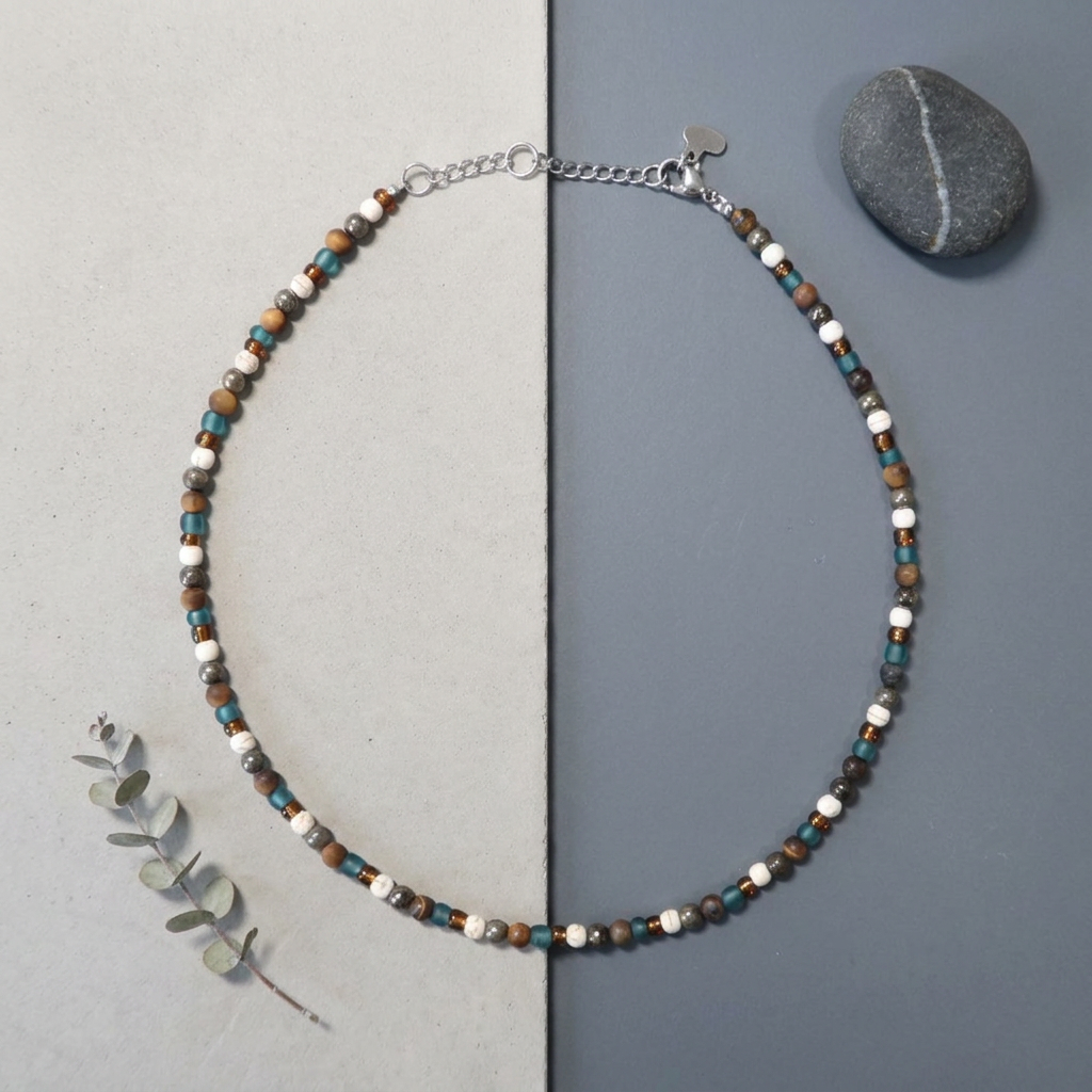 A beaded necklace with small brown, white, and turquoise-blue beads on a silver chain, displayed on a split gray background with a small stone and eucalyptus sprig. Tiger eyes, turquoise, pyrite and glass teal and amber.