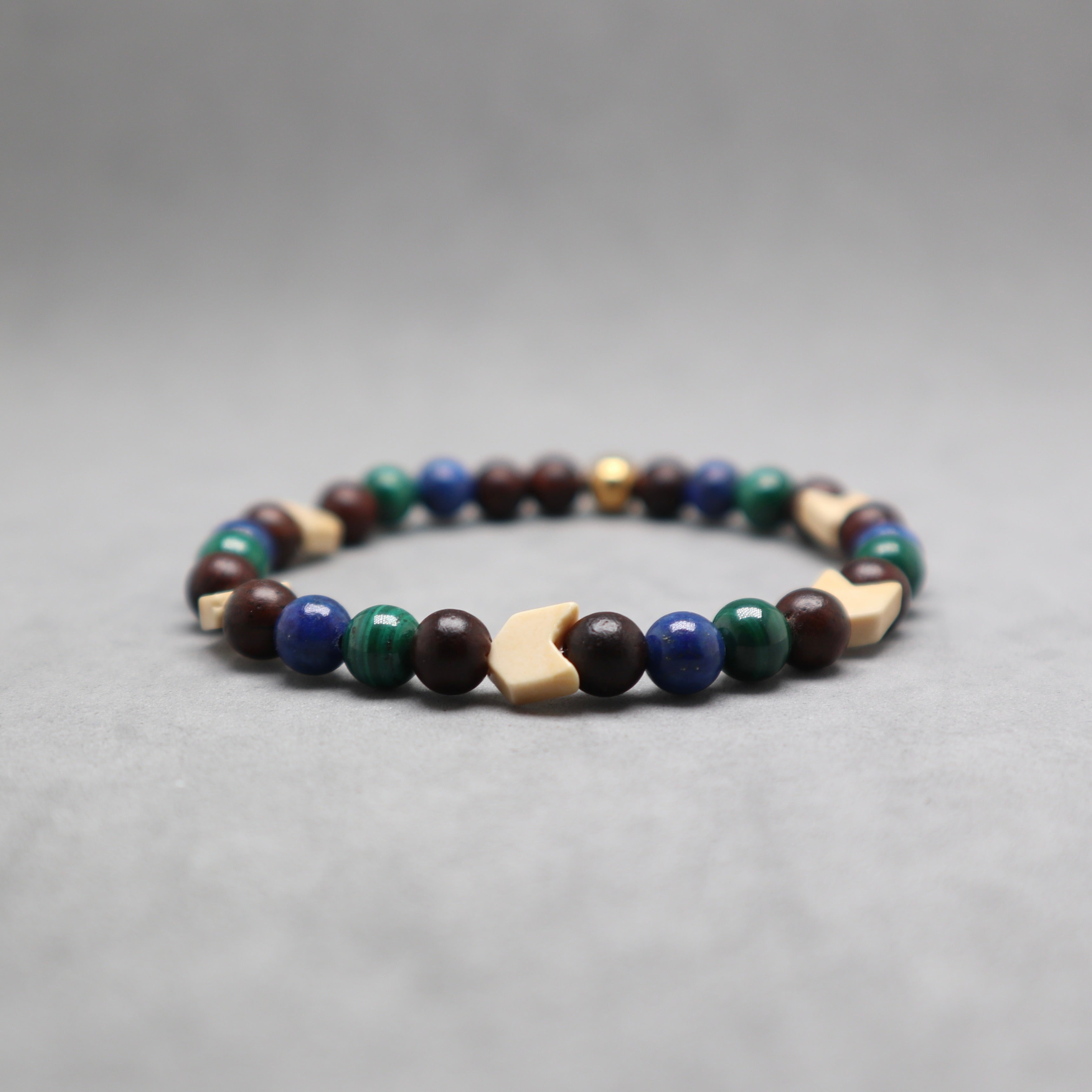 A beaded stretch bracelet featuring dark brown wooden beads, blue and green round beads, light beige geometric wooden beads, and a small metallic gold bead, displayed on a neutral gray background.