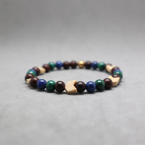 A beaded stretch bracelet featuring dark brown wooden beads, blue and green round beads, light beige geometric wooden beads, and a small metallic gold bead, displayed on a neutral gray background.