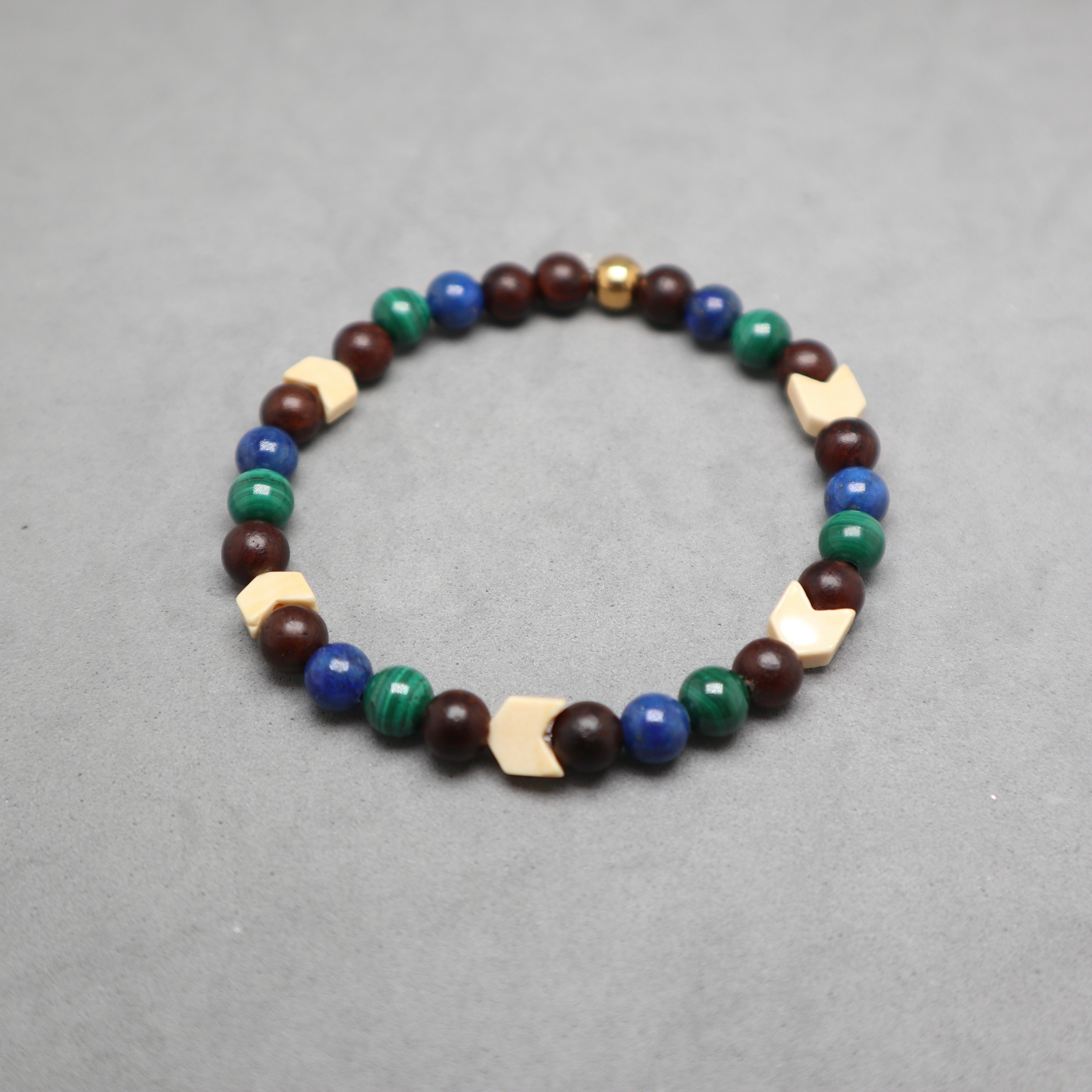 A beaded stretch bracelet featuring dark brown wooden beads, blue and green stone beads, and light beige geometric wooden accents, displayed on a neutral gray background. Lapis, malachite, snake agate and wood.