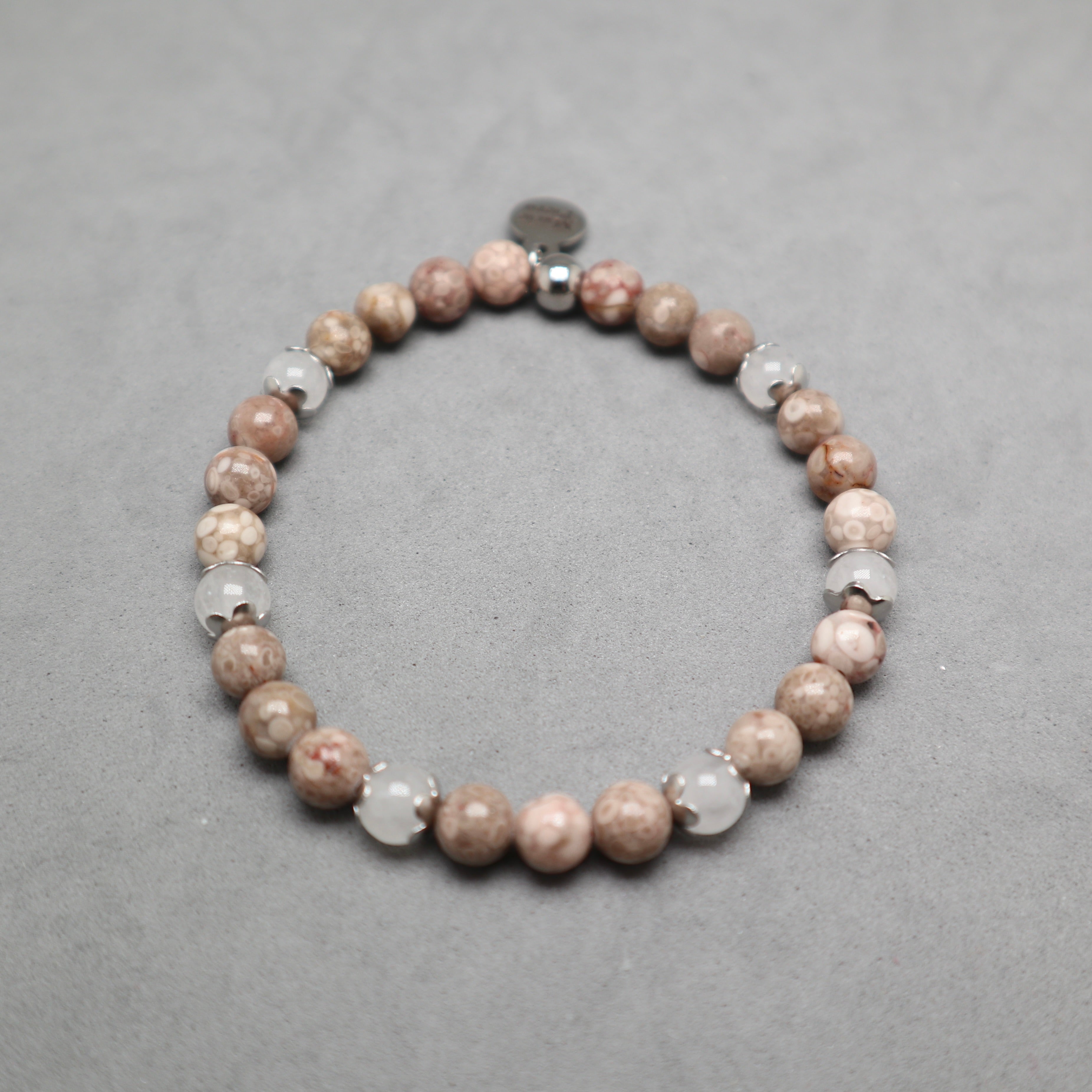 A beaded stretch bracelet featuring round brown and beige stone beads, clear faceted beads, and a small silver spacer, displayed on a textured gray background. Maifanite and jade