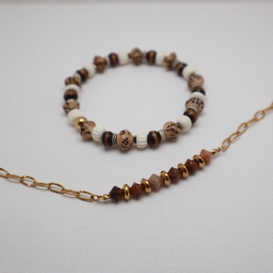 A beaded bracelet and matching beaded necklace with a gold chain, displayed on a light gray textured background. The beads feature natural brown, cream, and black marbled patterns, with some translucent pinkish-beige beads on the necklace.