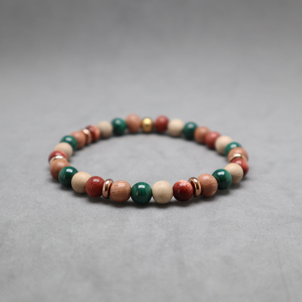 A beaded stretch bracelet made of round wooden beads in natural light brown, red, and green, accented with small metallic spacer rings, photographed on a neutral gray background.