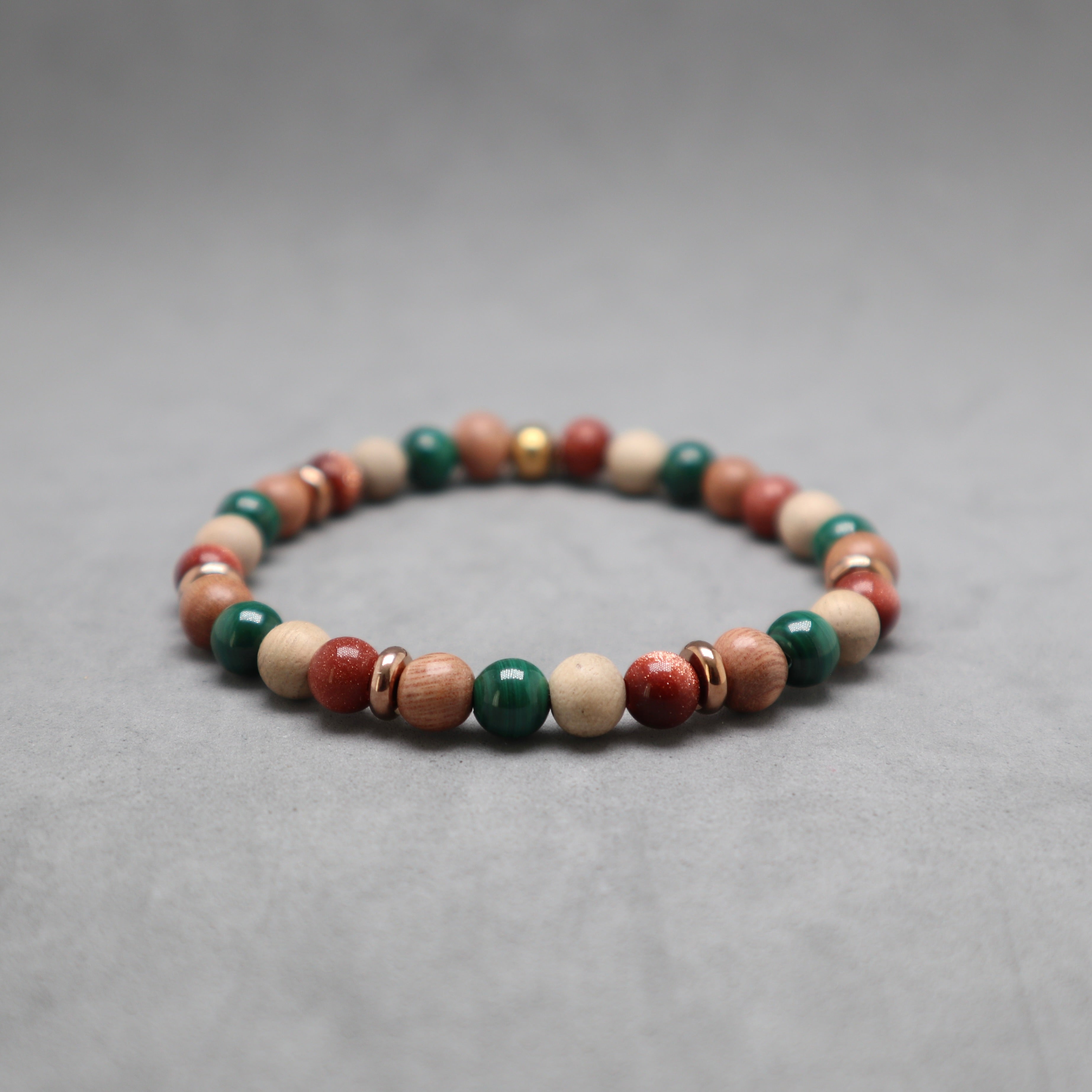 A beaded stretch bracelet made of round wooden beads in natural light brown, red, and green, accented with small metallic spacer rings, photographed on a neutral gray background.