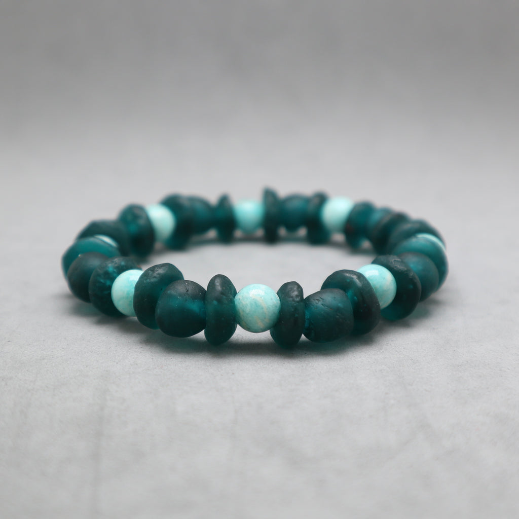 A beaded stretch bracelet made of dark teal-green and light aqua blue-green beads, photographed on a neutral gray background.
