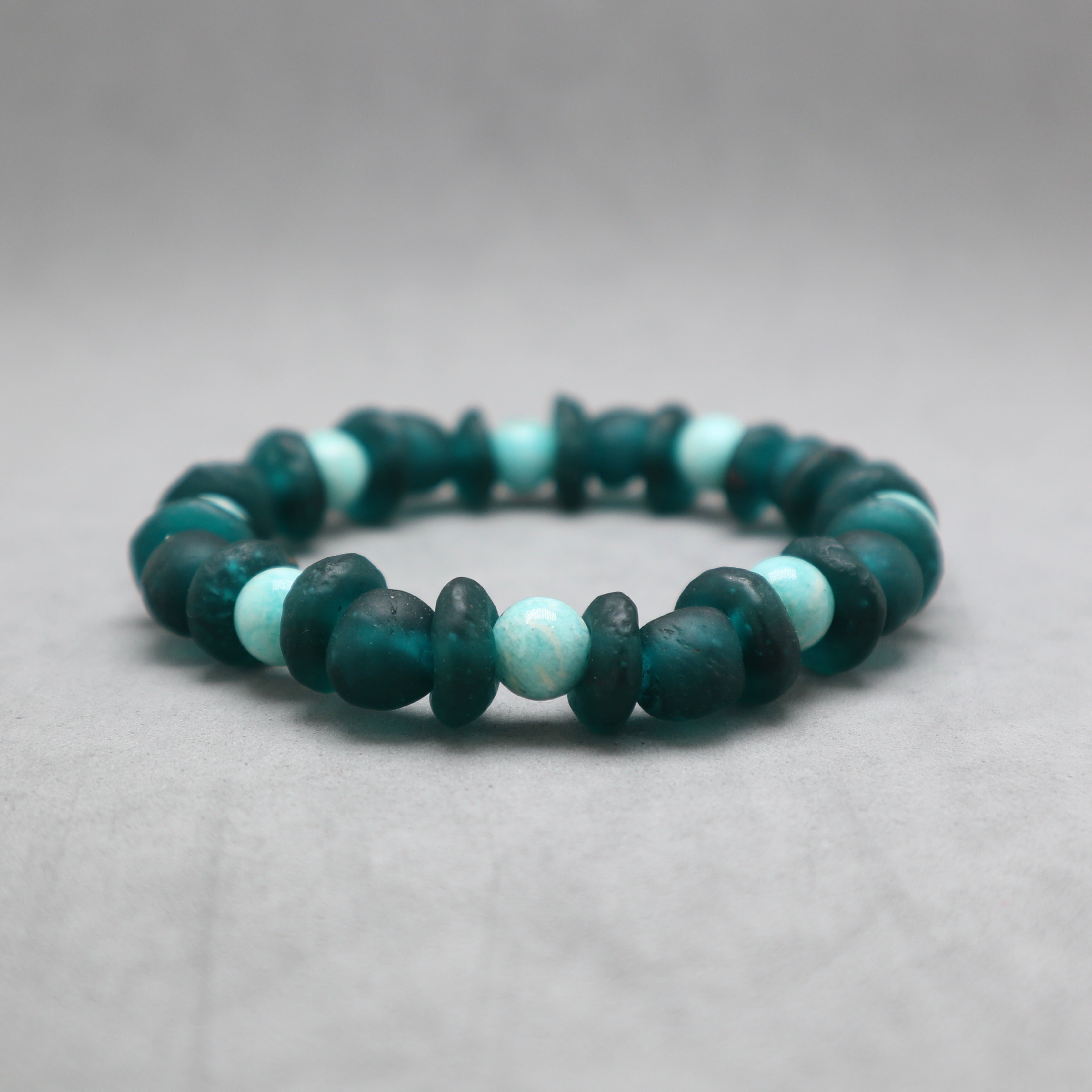A beaded stretch bracelet made of dark teal-green and light aqua blue-green beads, photographed on a neutral gray background.