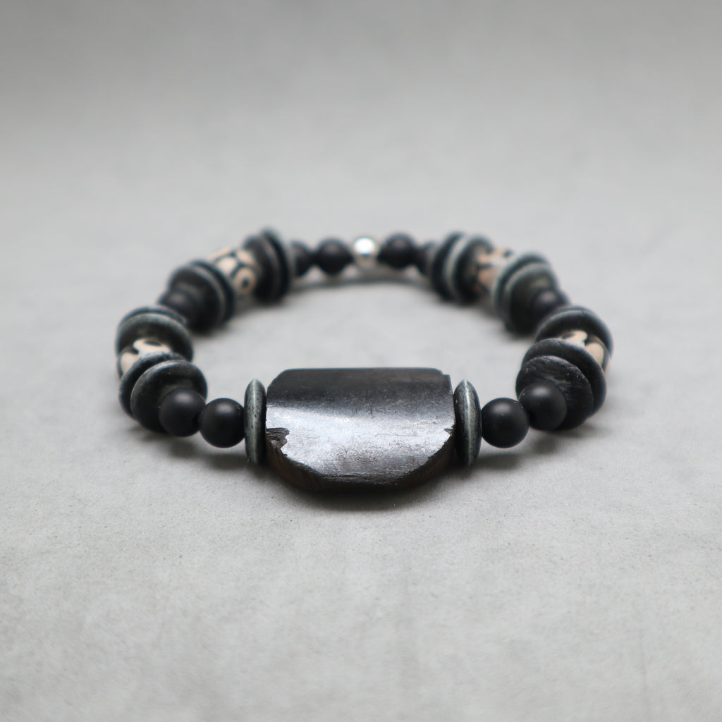 A beaded stretch bracelet featuring a large, polished dark gray central stone surrounded by matte black beads and patterned spacer beads, set against a neutral gray background.