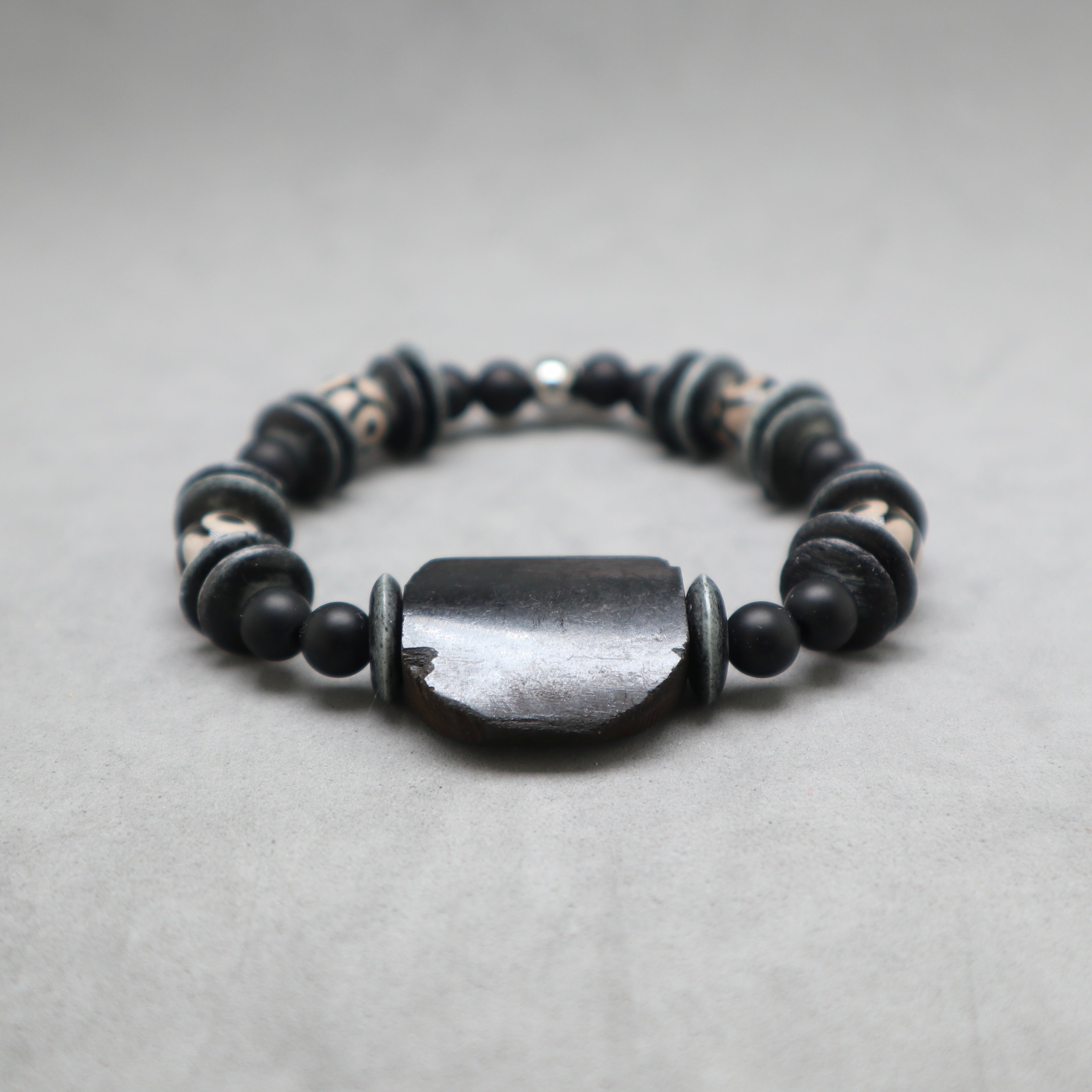 A beaded stretch bracelet featuring a large, polished dark gray central stone surrounded by matte black beads and patterned spacer beads, set against a neutral gray background.