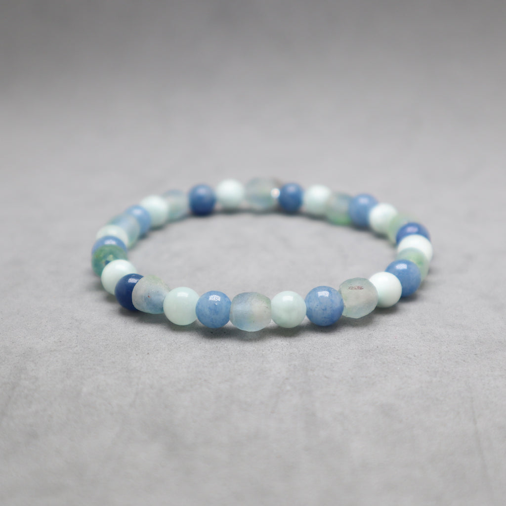 A beaded stretch bracelet made of round polished gemstone beads in shades of light blue, medium blue, white, and pale mint green, displayed on a neutral gray textured surface.