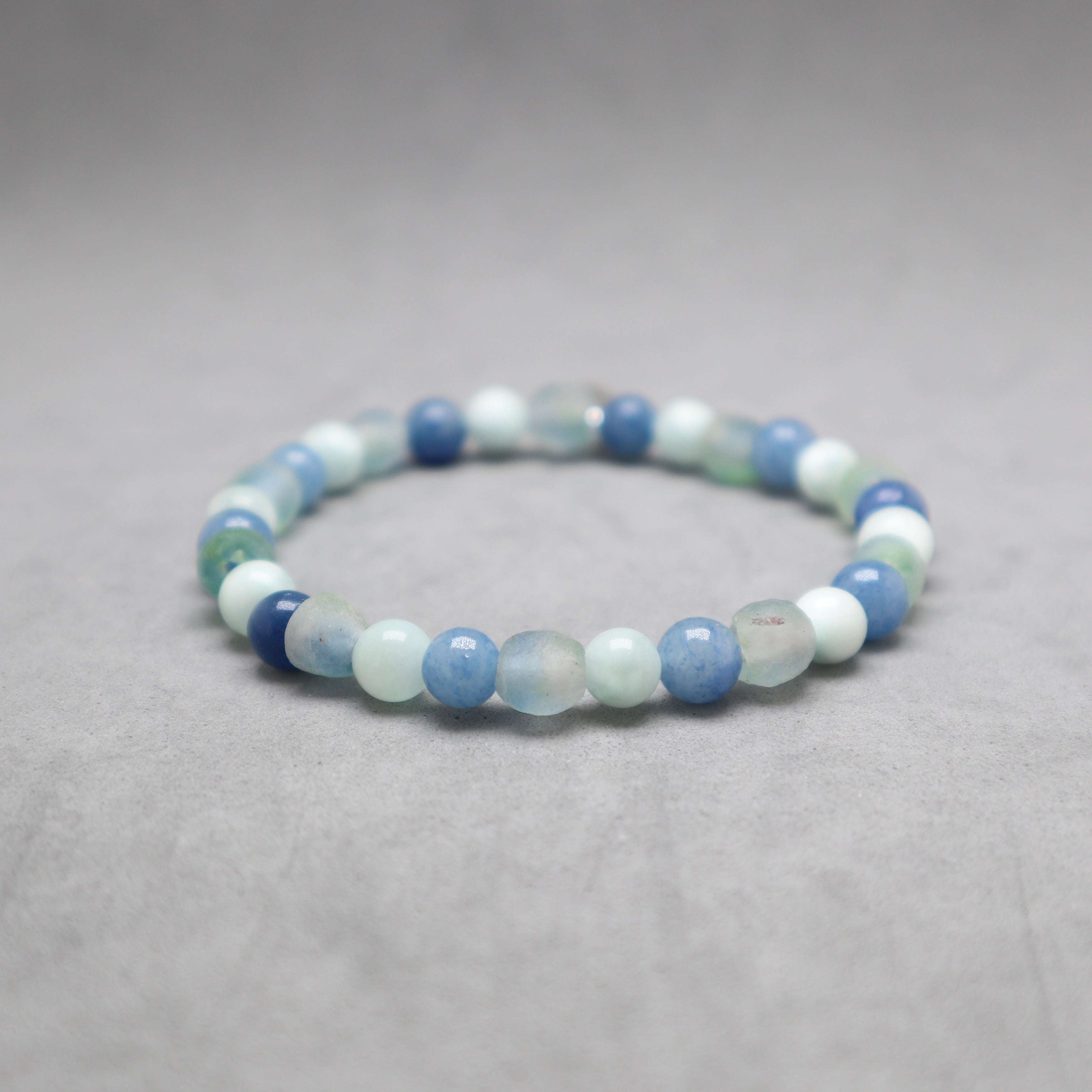 A beaded stretch bracelet made of round polished gemstone beads in shades of light blue, medium blue, white, and pale mint green, displayed on a neutral gray textured surface.
