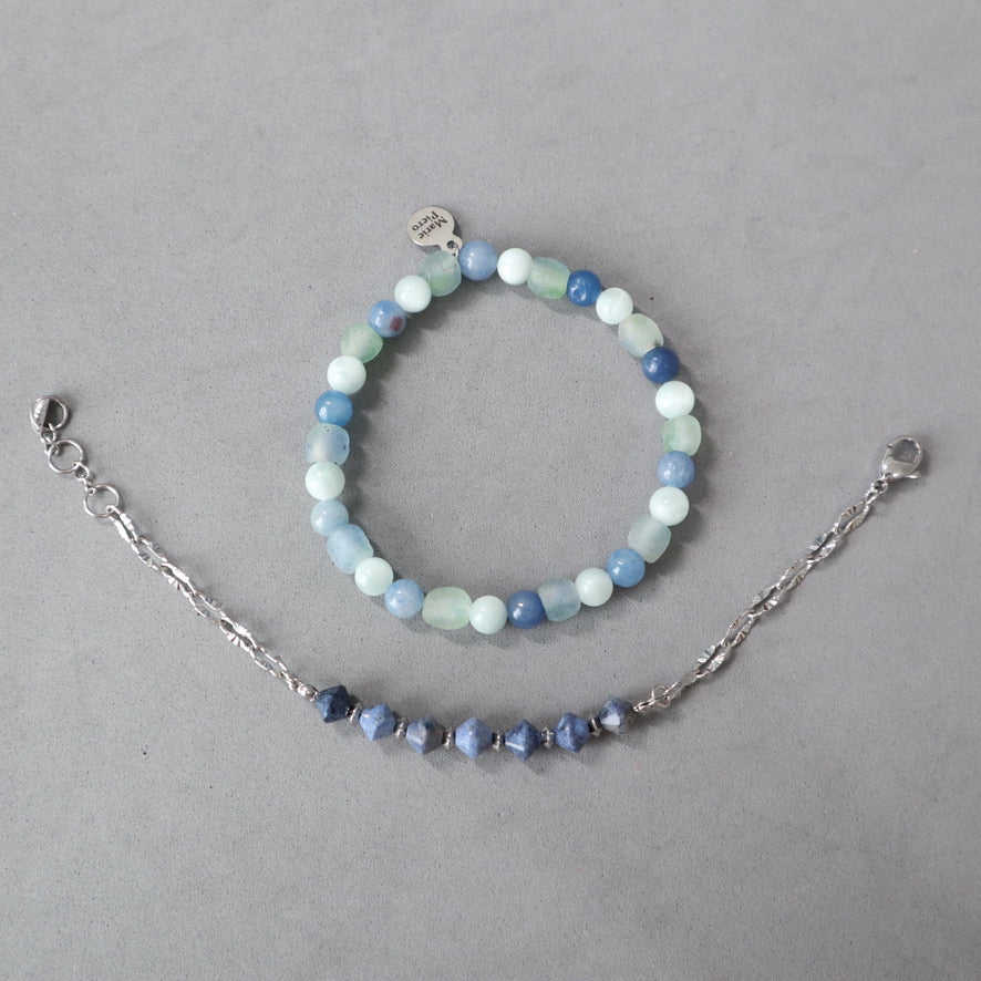 A delicate beaded bracelet and matching chain bracelet laid flat on a neutral gray background. The beaded bracelet features round beads in shades of light blue, medium blue, and pale mint green, with a small silver charm.