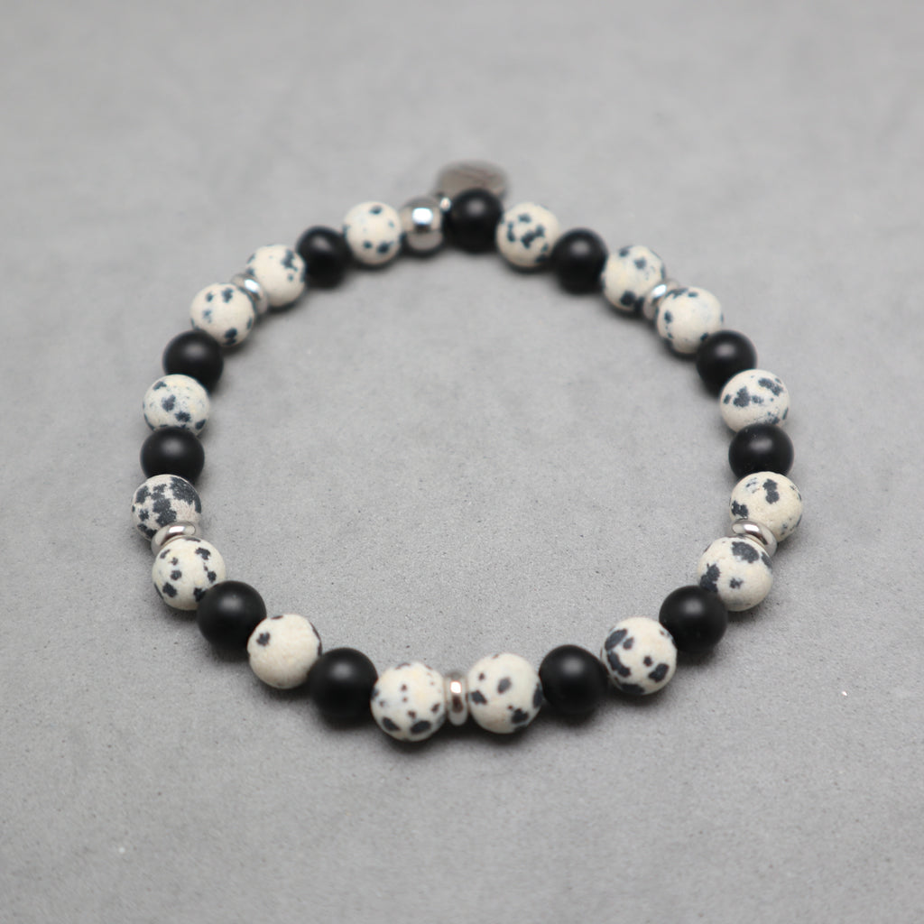 A beaded stretch bracelet featuring alternating matte black beads and speckled white and black stone beads, displayed on a neutral gray textured surface.