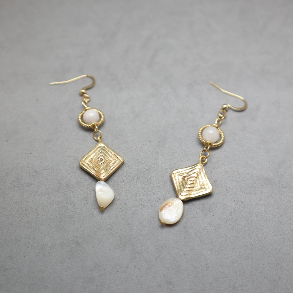 A pair of gold dangle earrings with textured geometric charms, small white shell beads, and irregular white shell drops, displayed on a neutral gray background.