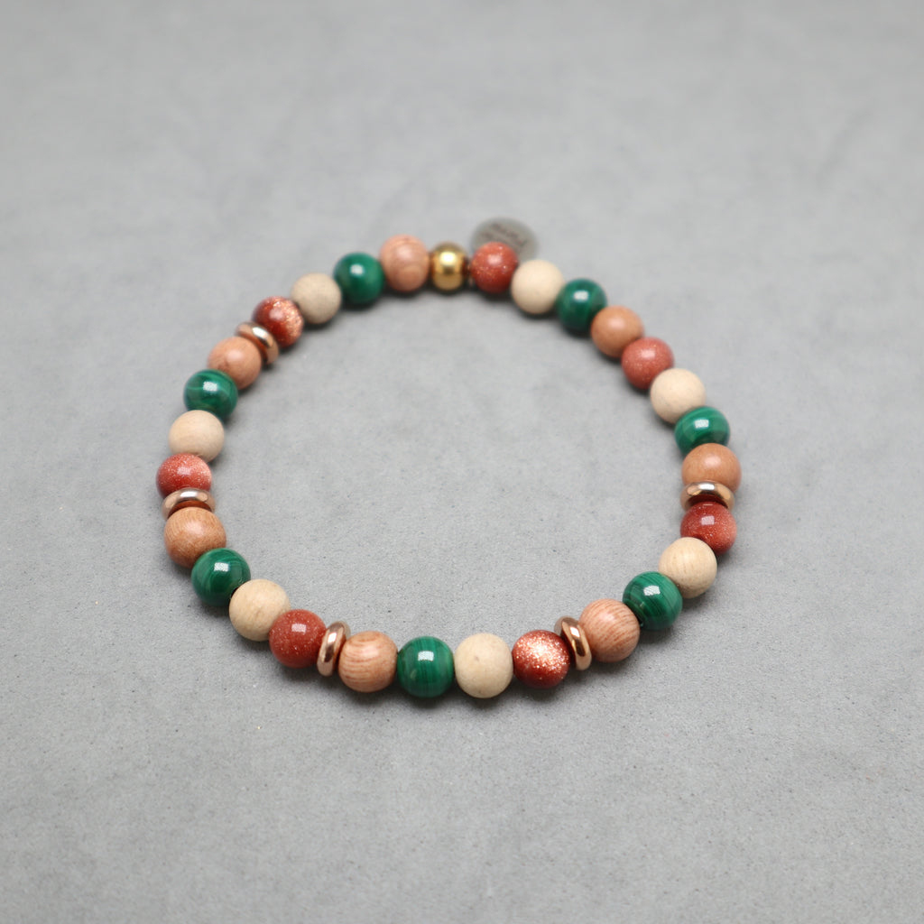A beaded stretch bracelet featuring natural wood beads, green malachite beads, red coral beads, and small metallic spacer beads, displayed on a neutral gray background.