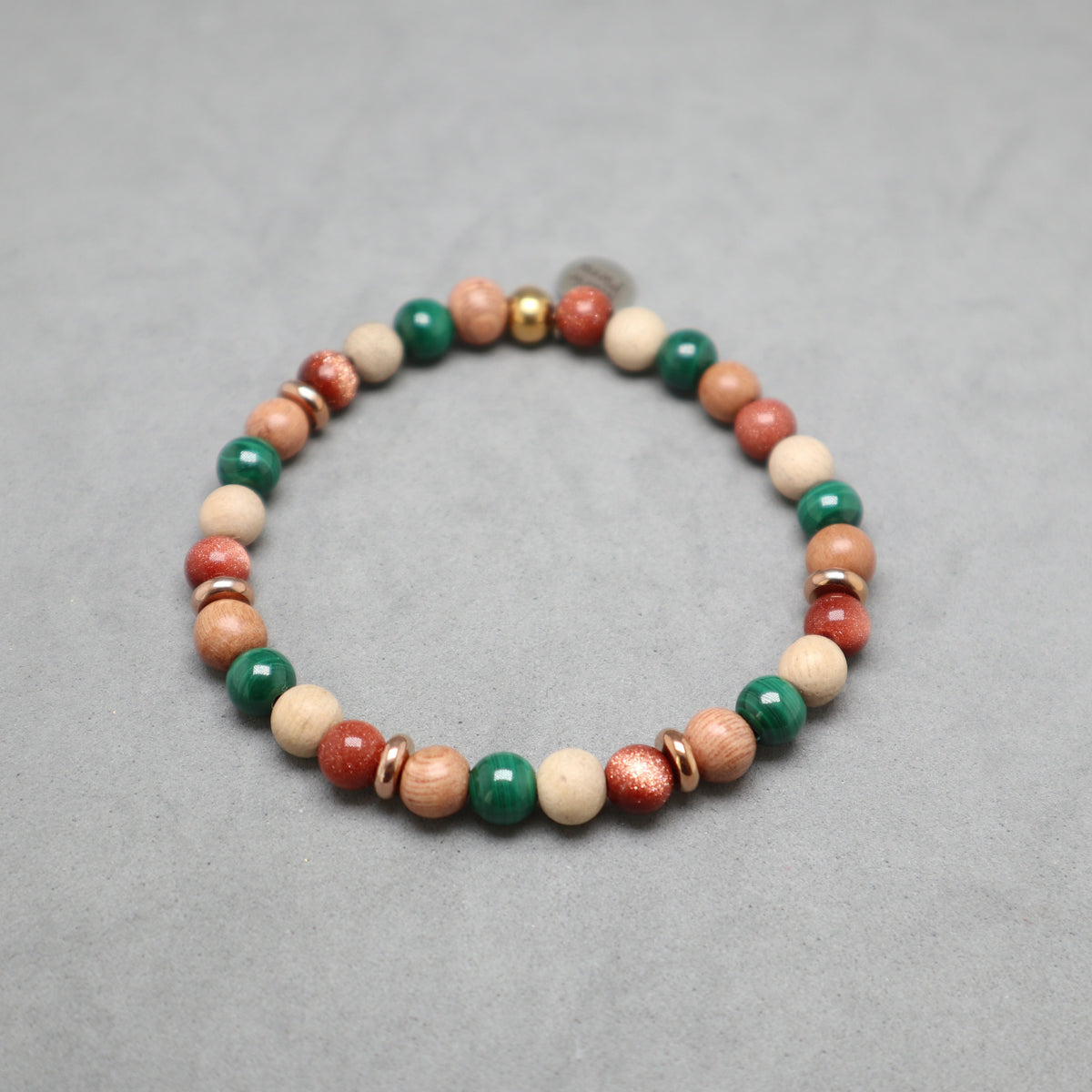 A beaded stretch bracelet featuring natural wood beads, green malachite beads, red coral beads, and small metallic spacer beads, displayed on a neutral gray background.