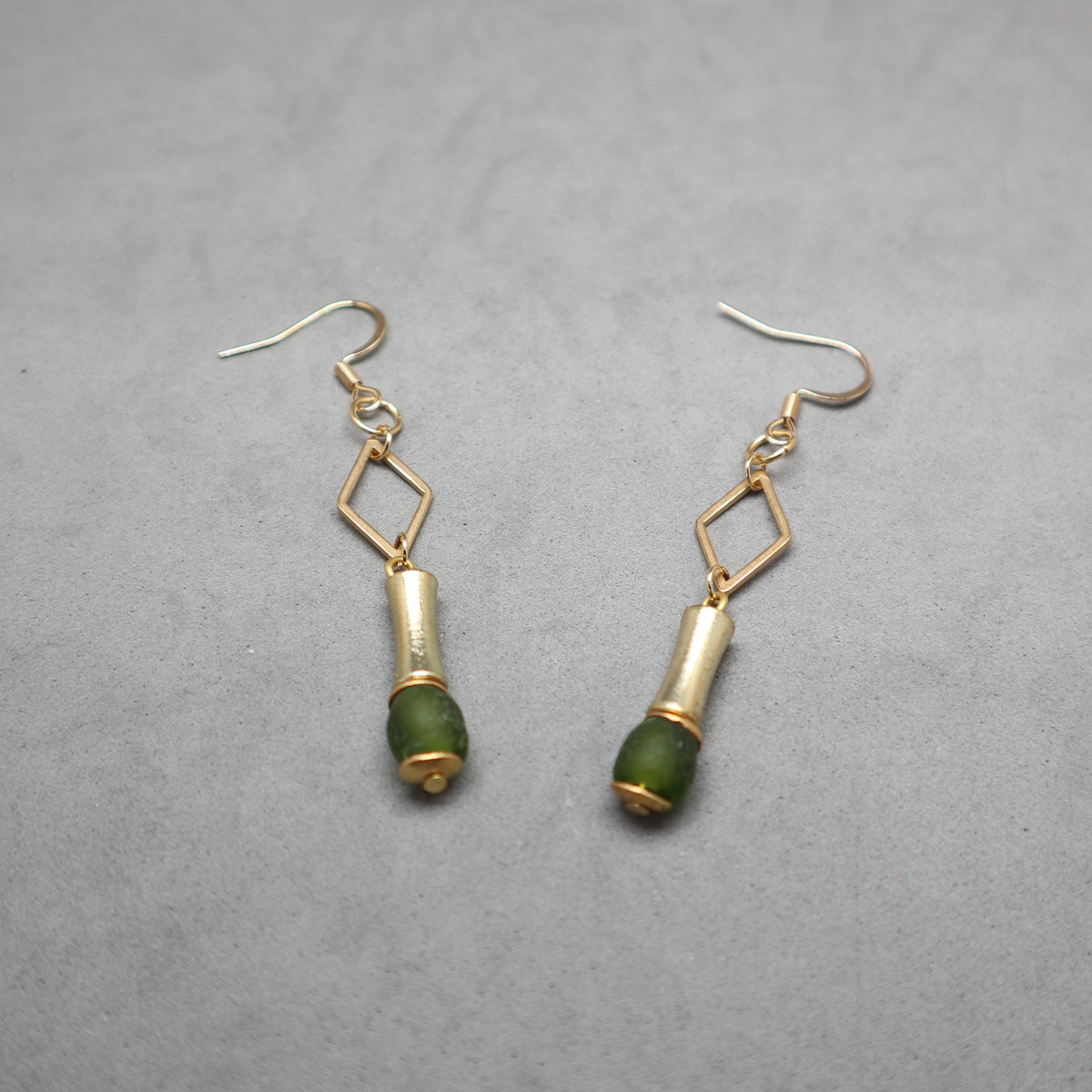 A pair of gold and green drop earrings with geometric diamond-shaped links and cylindrical green bead drops, displayed on a textured gray background.