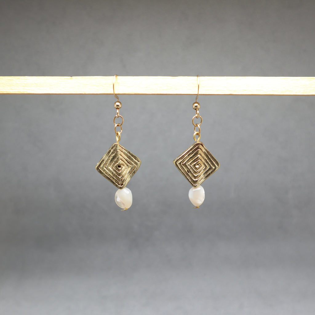 A pair of gold-tone geometric drop earrings with a textured square pendant and a small white pearl dangle, hanging from a light wooden display bar against a neutral gray background.