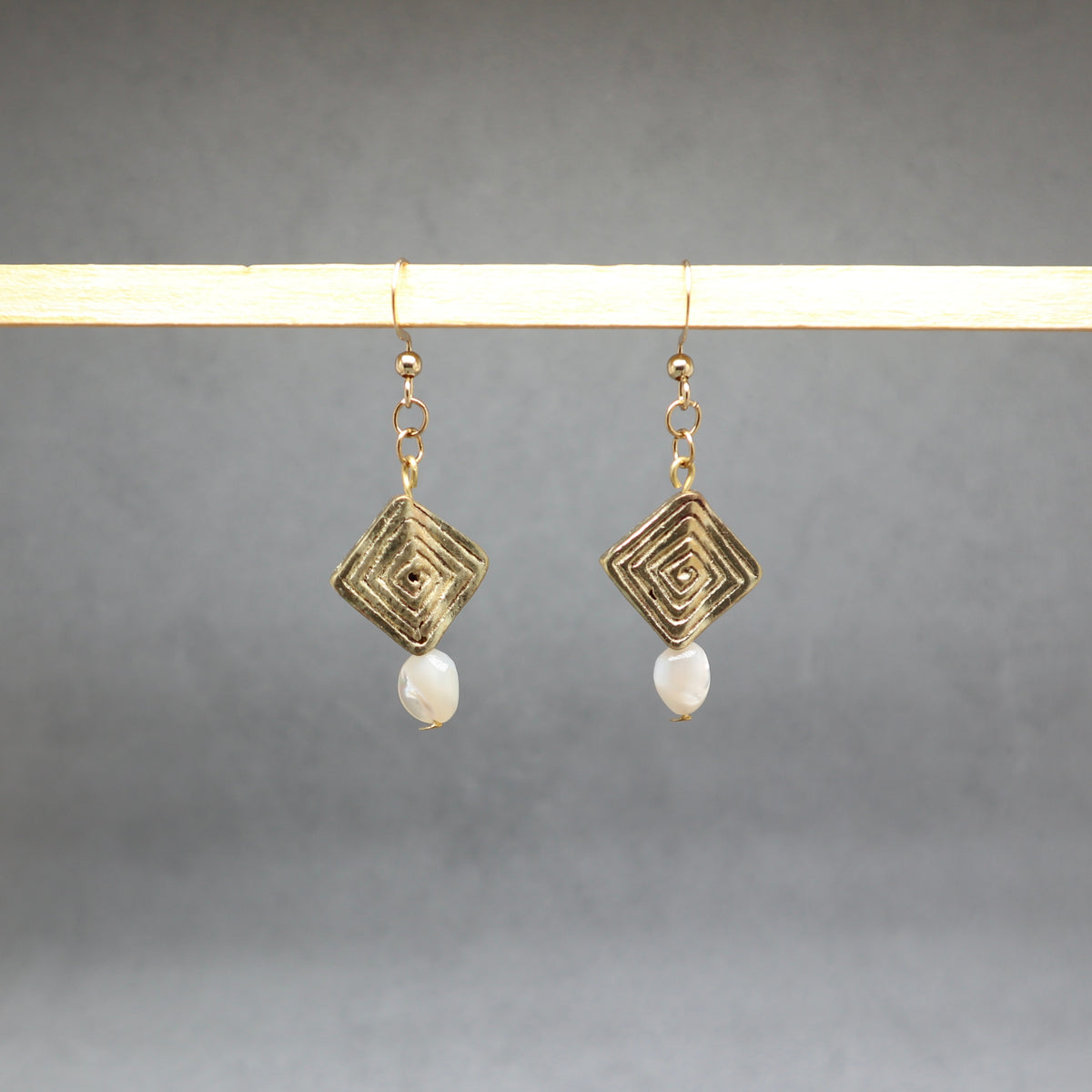 A pair of gold-tone geometric drop earrings with a textured square pendant and a small white pearl dangle, hanging from a light wooden display bar against a neutral gray background.