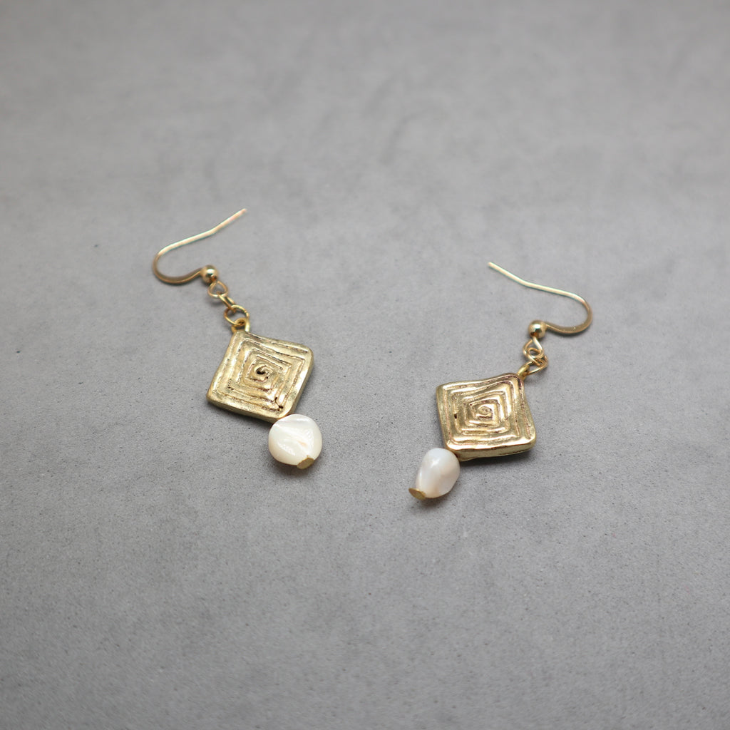A pair of gold-toned dangle earrings with textured square charms and small white shell-like beads, displayed on a neutral gray background.