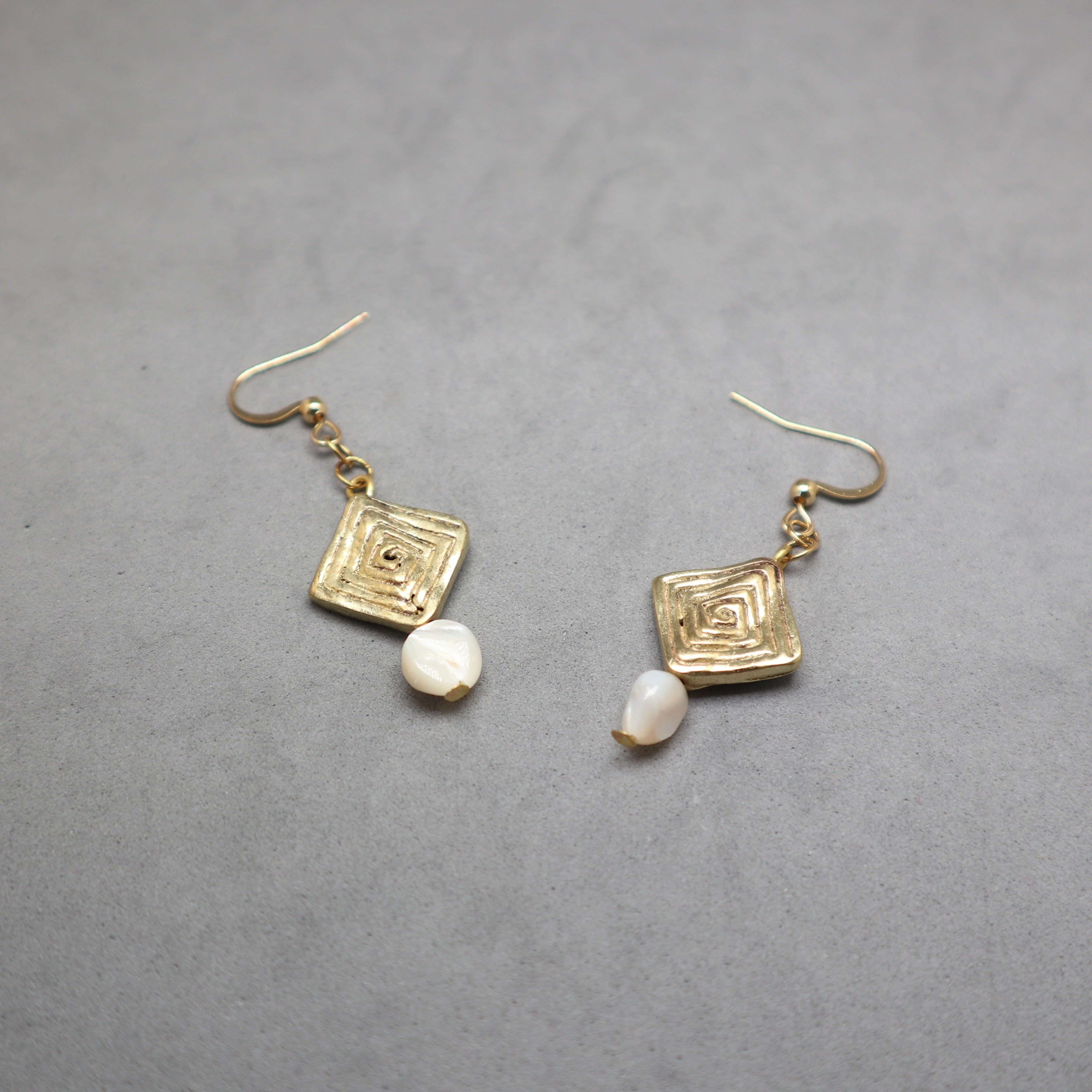 A pair of gold-toned dangle earrings with textured square charms and small white shell-like beads, displayed on a neutral gray background.