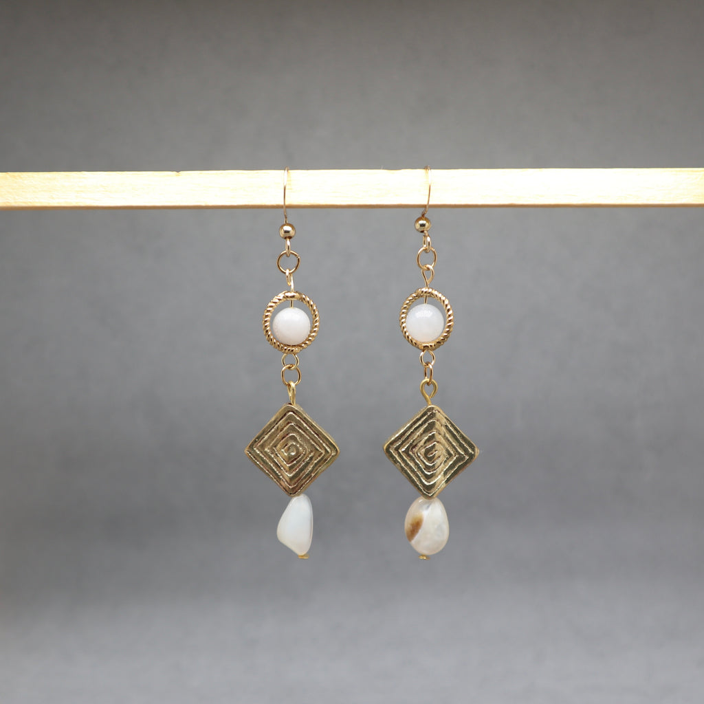 A pair of gold-toned dangle earrings with circular white bead details, geometric wirework diamond charms, and irregular white shell drops, hanging from a light wooden bar against a neutral gray background.