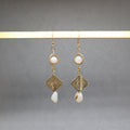 A pair of gold-toned dangle earrings with circular white bead details, geometric wirework diamond charms, and irregular white shell drops, hanging from a light wooden bar against a neutral gray background.