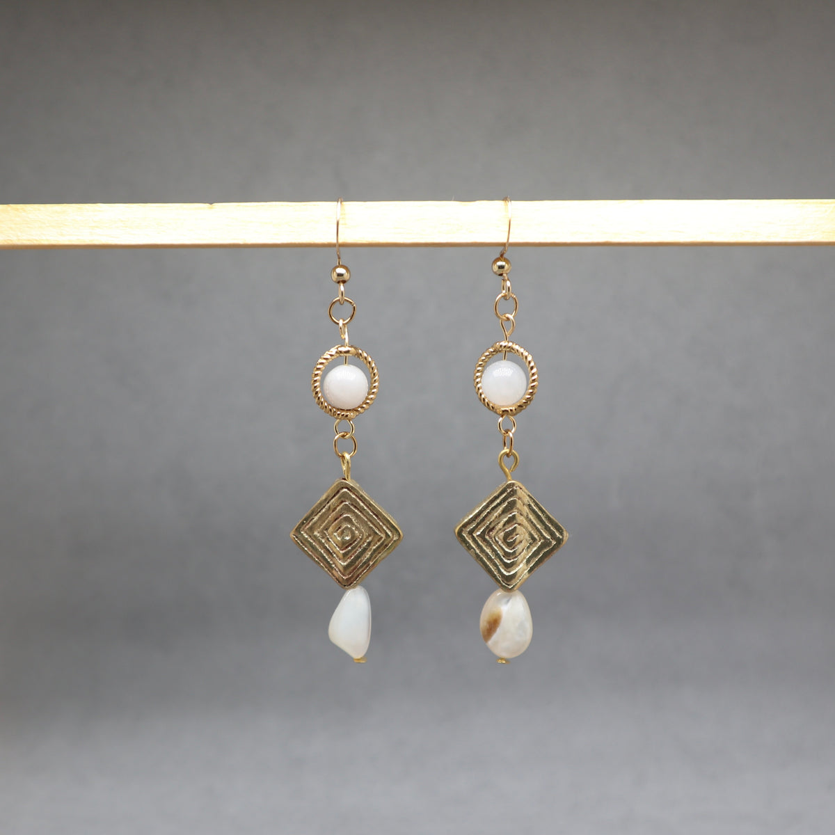 A pair of gold-toned dangle earrings with circular white bead details, geometric wirework diamond charms, and irregular white shell drops, hanging from a light wooden bar against a neutral gray background.