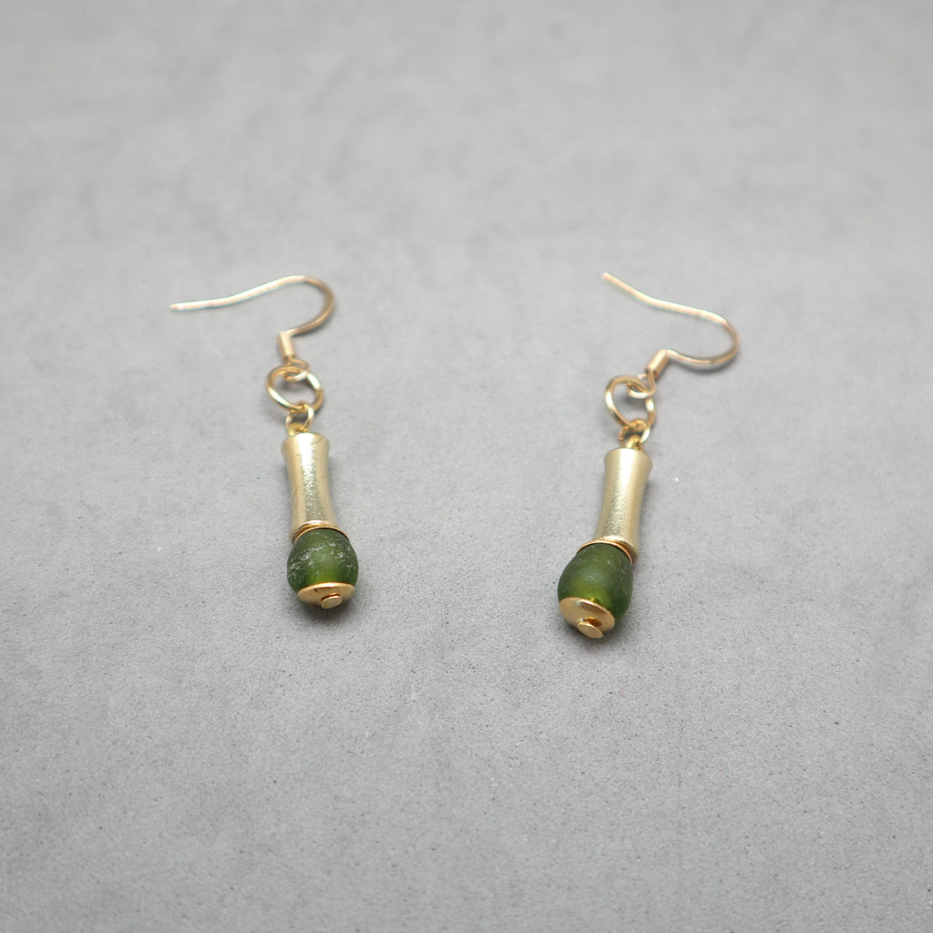 A pair of gold-tone dangle earrings with small cylindrical gold beads and round green stone beads, shown on a textured gray background.