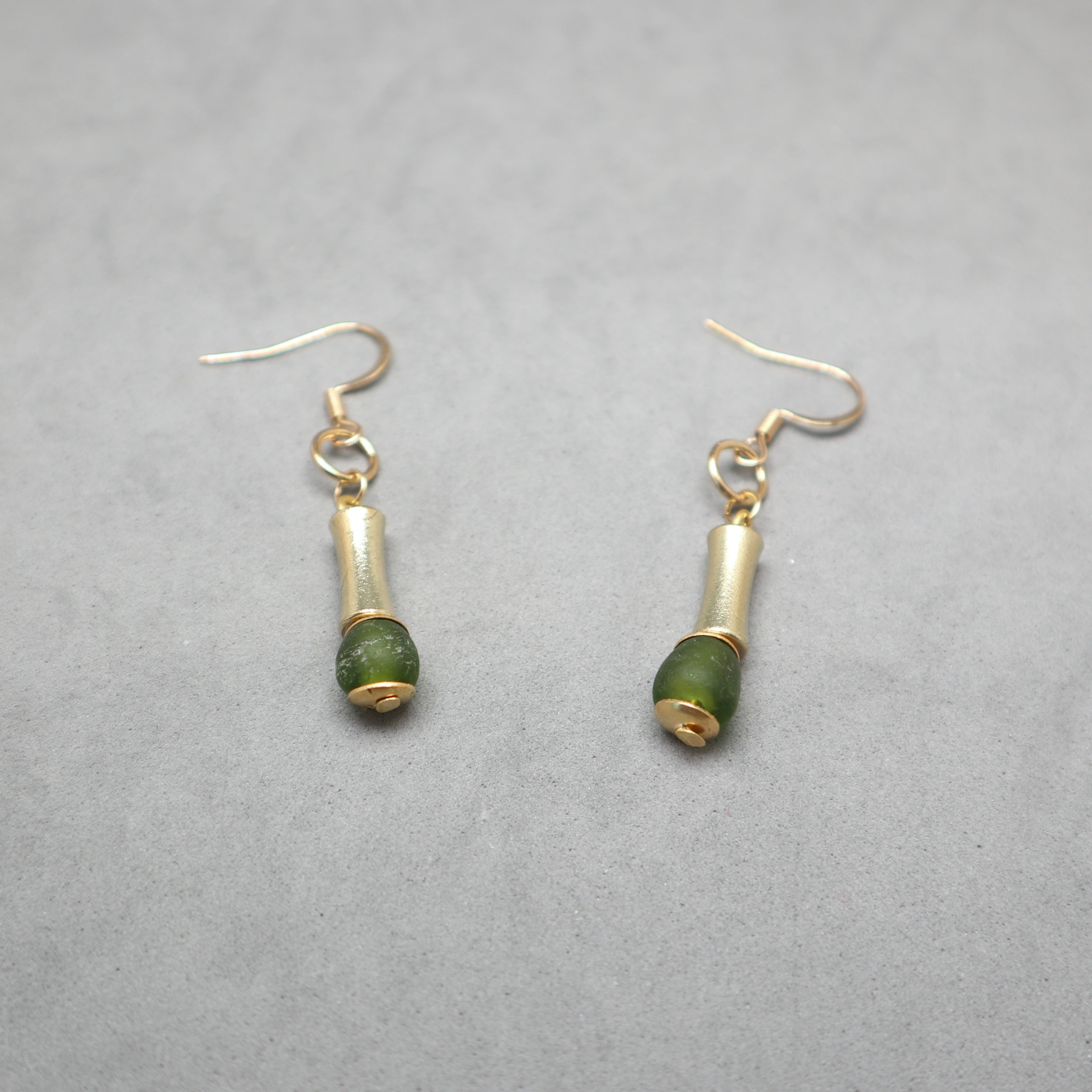 A pair of gold-tone dangle earrings with small cylindrical gold beads and round green stone beads, shown on a textured gray background.