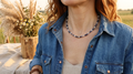 A close-up lifestyle shot of a woman wearing a beaded necklace with blue, white, and gray beads, styled with a denim shirt in an outdoor setting. Jave stripe beads, sodalite, jade, shell, metal heishi.