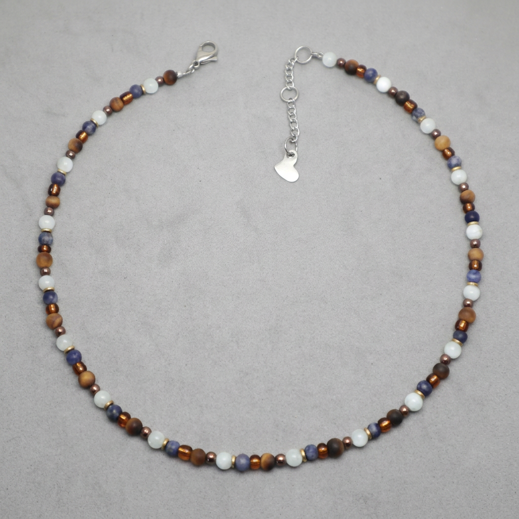 A beaded necklace with alternating white, brown, and blue-gray beads, featuring a silver lobster clasp and a silver heart-shaped charm on an elastic chain, displayed on a neutral gray background.