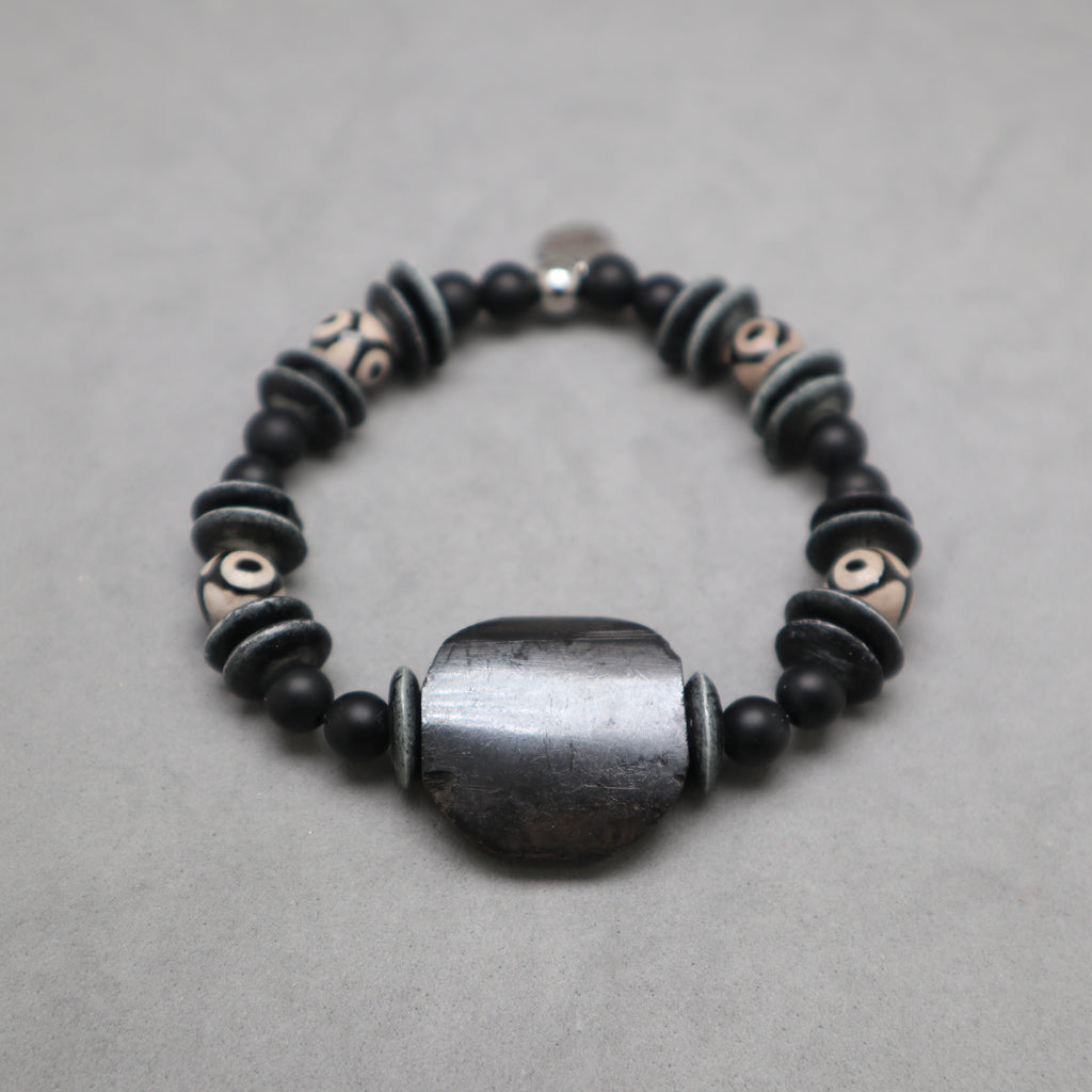 A beaded stretch bracelet featuring matte black round beads, textured dark gray disc beads with white symbols, and a large central dark gray stone bead, displayed on a neutral gray background. Kenya and himalayan bone, black onyx, eye agate