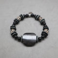 A beaded stretch bracelet featuring matte black round beads, textured dark gray disc beads with white symbols, and a large central dark gray stone bead, displayed on a neutral gray background. Kenya and himalayan bone, black onyx, eye agate