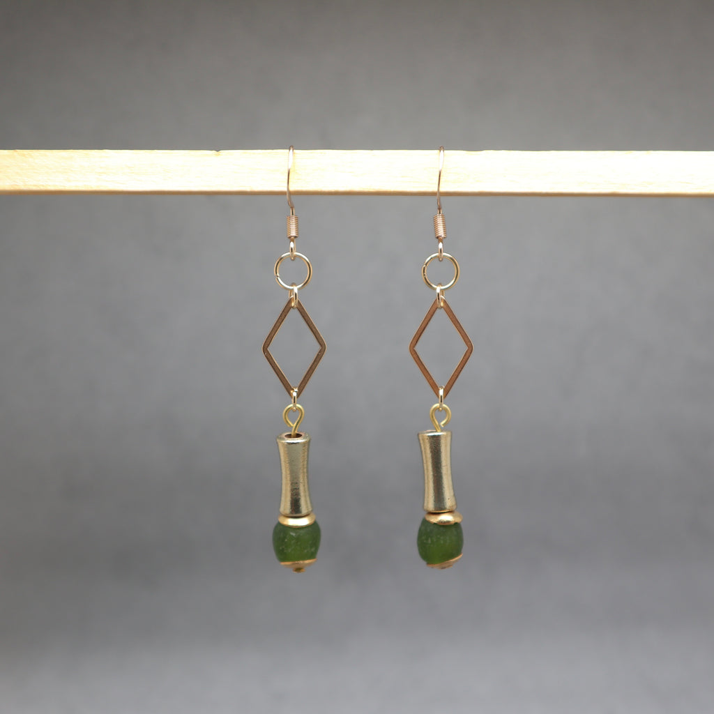 A pair of dangling earrings hanging from a wooden display bar against a soft gray background. Each earring has a gold-tone hook and diamond-shaped frame, a silver-tone cylindrical drop, and a green faceted bead at the bottom.