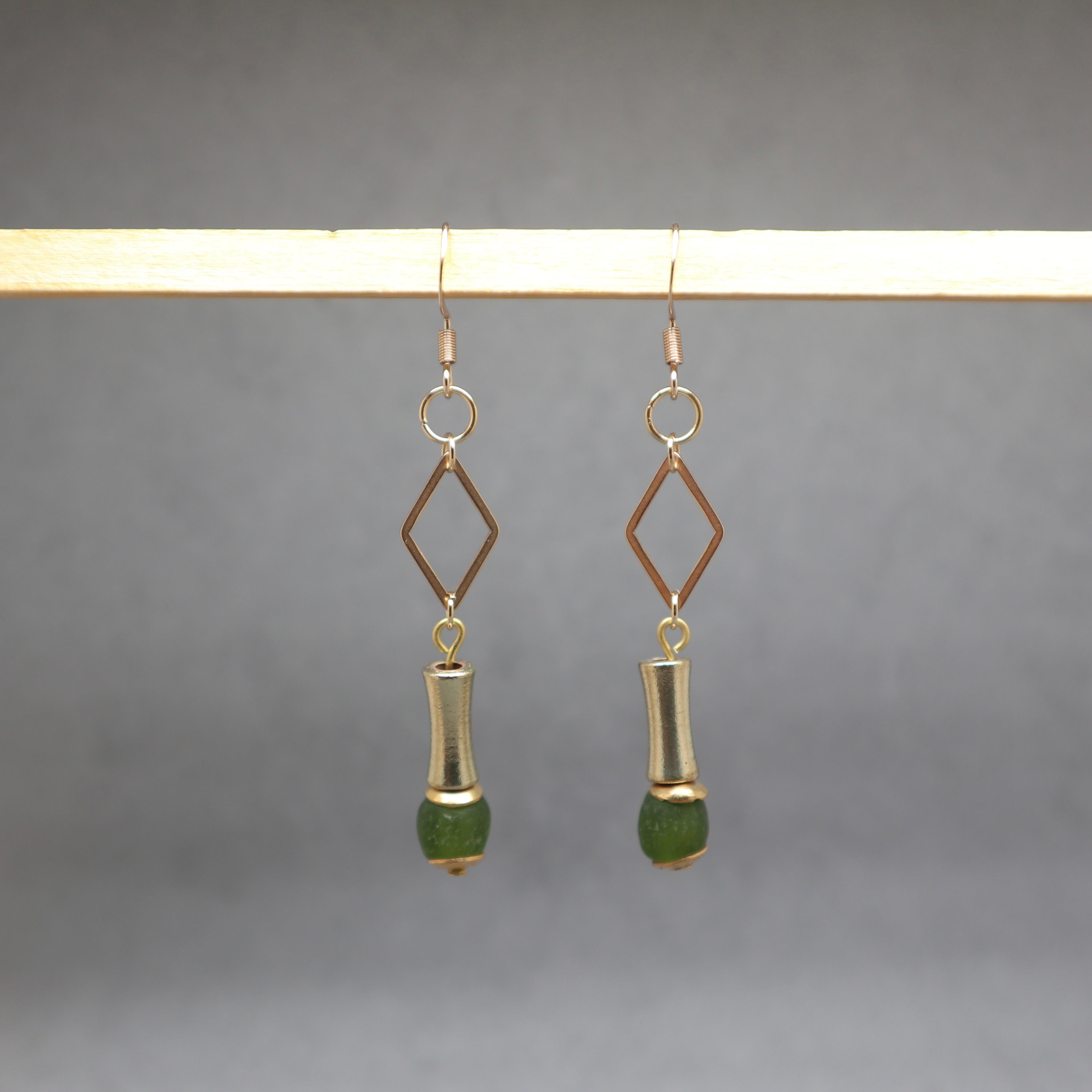 A pair of dangling earrings hanging from a wooden display bar against a soft gray background. Each earring has a gold-tone hook and diamond-shaped frame, a silver-tone cylindrical drop, and a green faceted bead at the bottom.