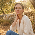 A woman wearing a cream linen long-sleeve top and blue jeans sits outdoors in a sunny, sunlit environment with trees and a stone wall in the background.