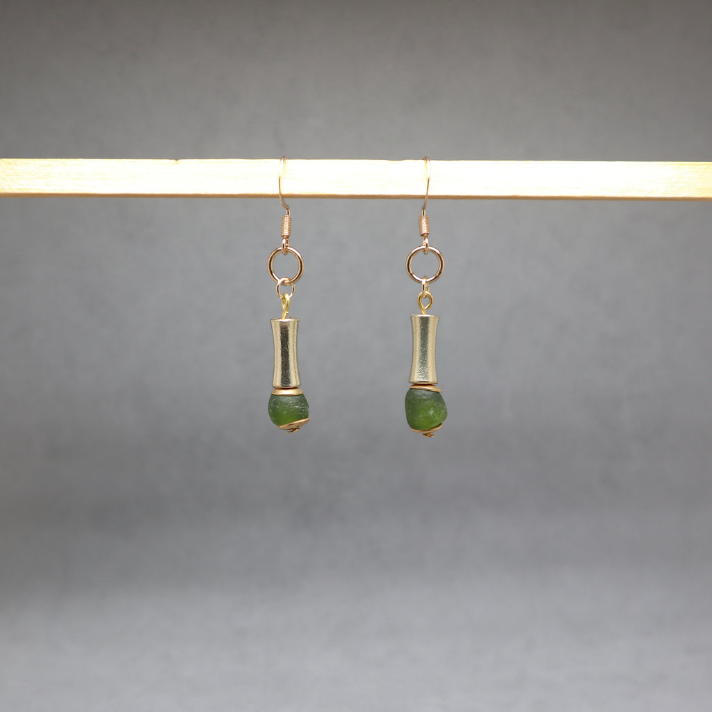 A pair of gold-toned dangle earrings with cylindrical metal links and small green bead drops, hanging from a light wooden display bar against a soft gray background.