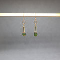 A pair of gold-toned dangle earrings with cylindrical metal links and small green bead drops, hanging from a light wooden display bar against a soft gray background.