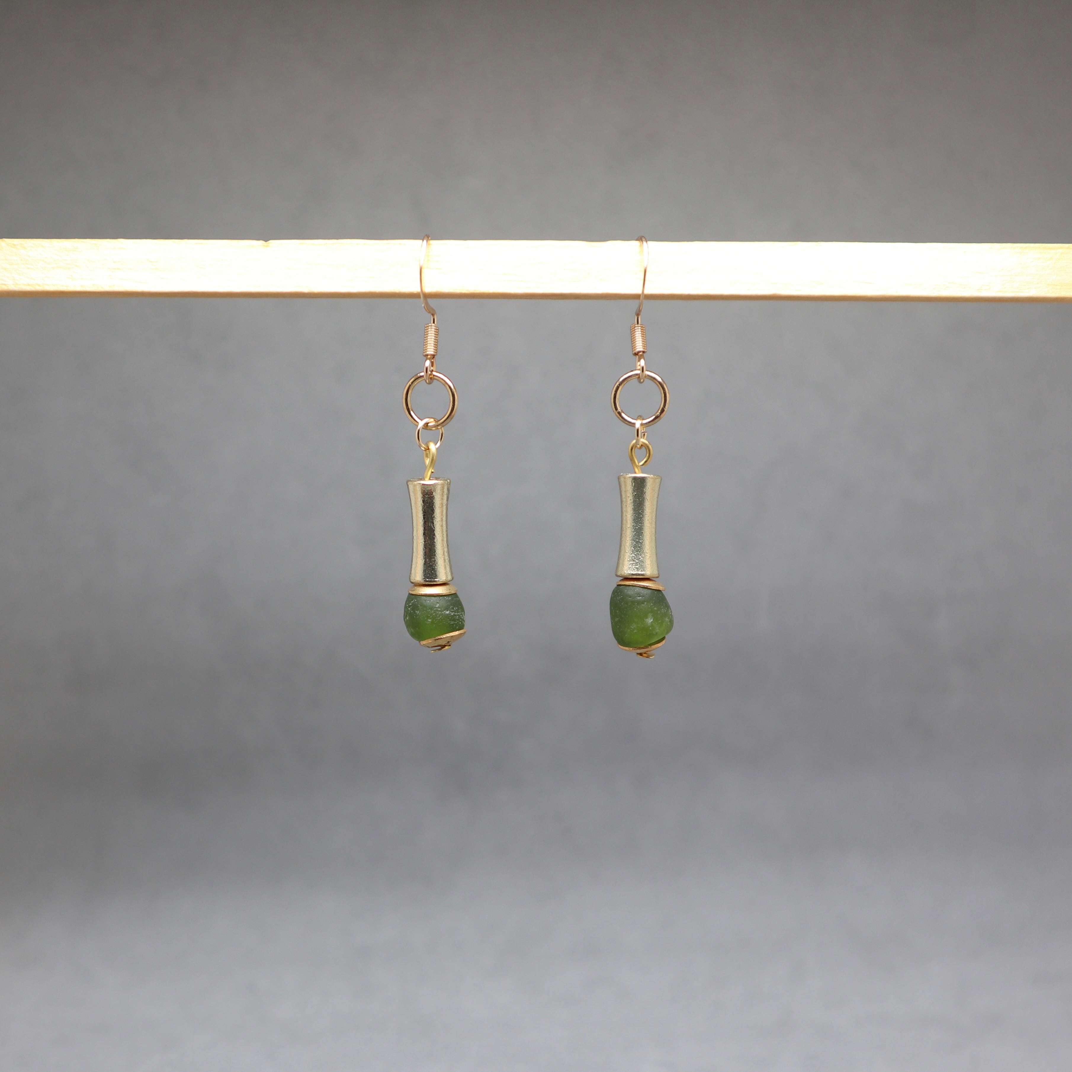 A pair of gold-toned dangle earrings with cylindrical metal links and small green bead drops, hanging from a light wooden display bar against a soft gray background.