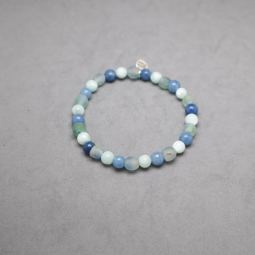 A beaded stretch bracelet made of round gemstone beads in shades of light blue, medium blue, white, and green, displayed on a neutral gray background.