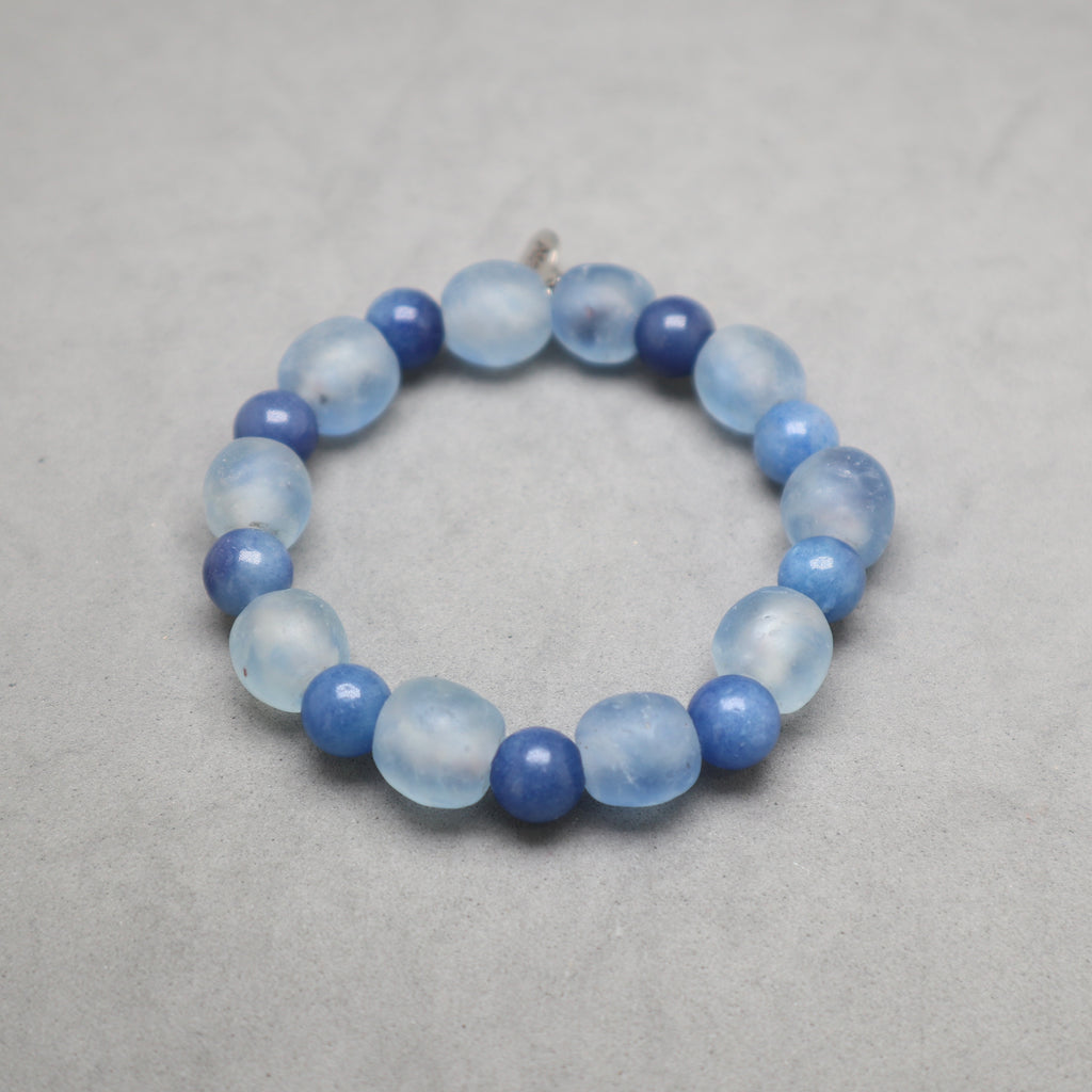 A beaded stretch bracelet made of light blue and medium blue translucent and opaque round beads, displayed on a neutral gray textured surface.