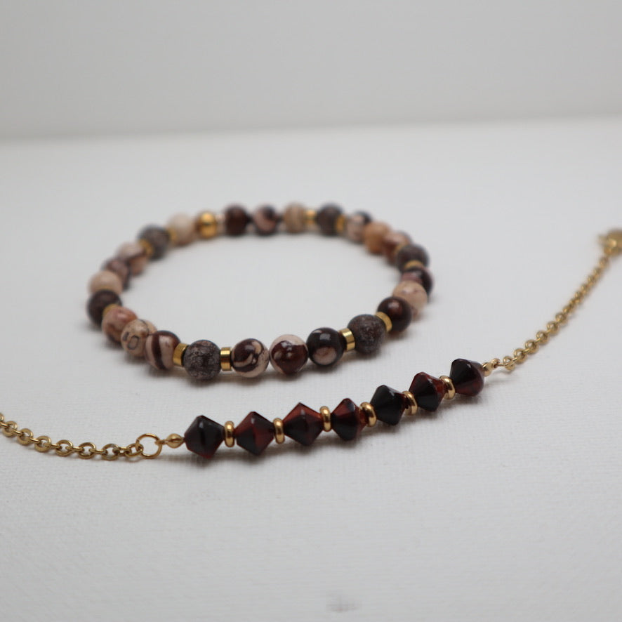 A beaded bracelet and matching chain necklace displayed on a light gray surface. The jewelry features marbled beads in brown, beige, and dark tones, accented with gold spacers and a gold chain with faceted dark red beads.
